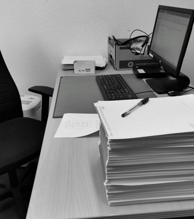 Black and white image of a cluttered office desk with a computer, chair, and paperwork.